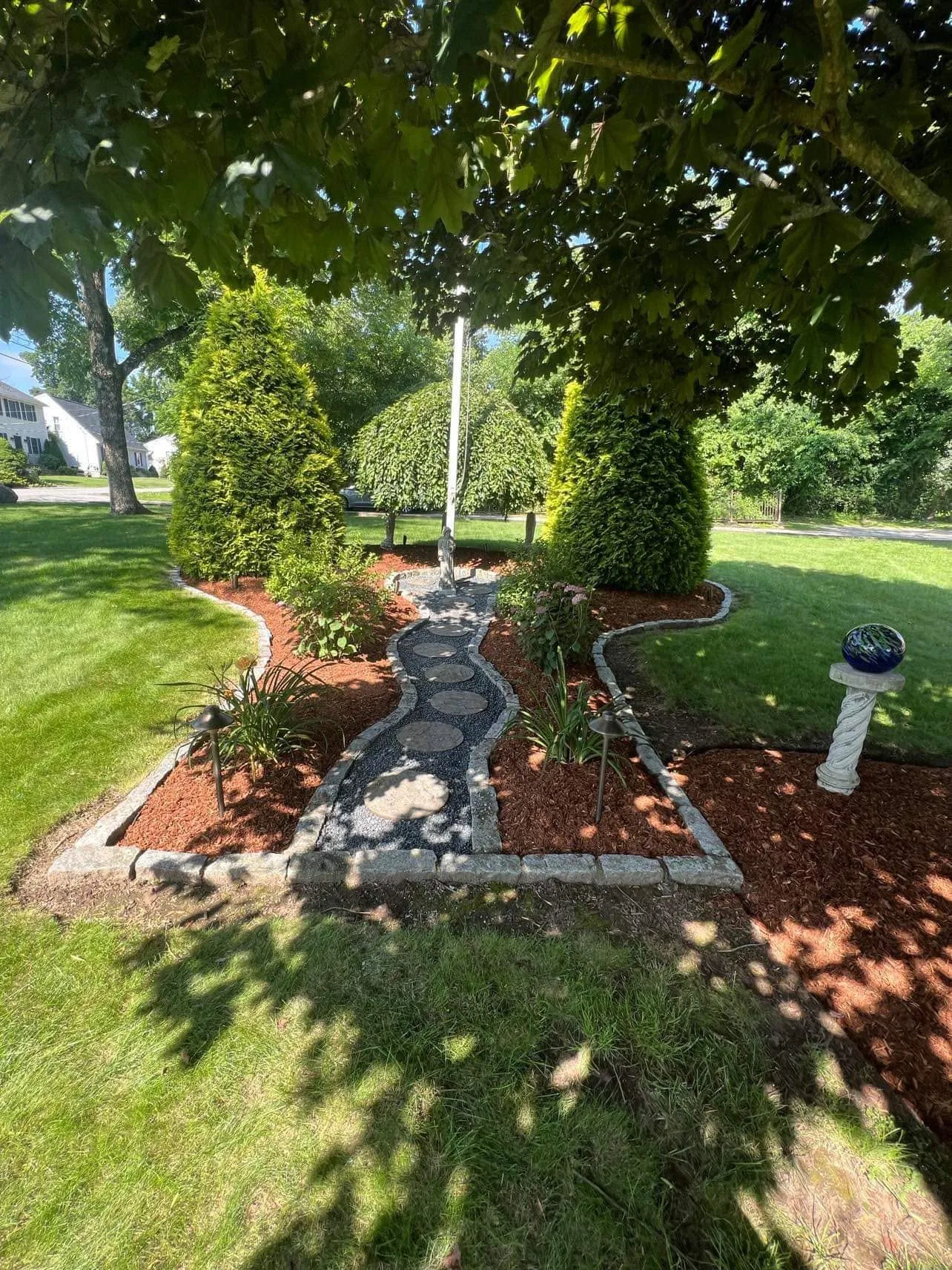 Mulched garden bed with a stone path and a birdbath under a tree